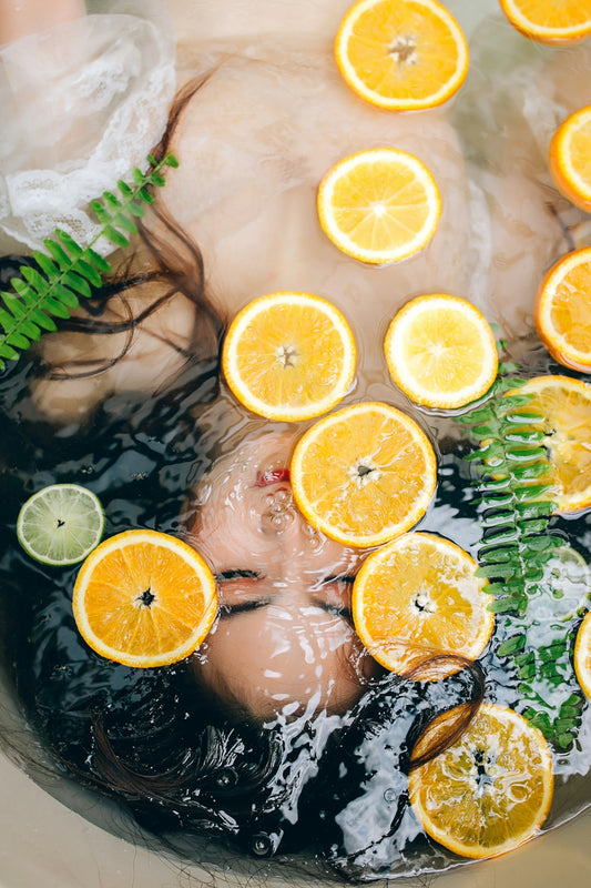 A person submerged in water with floating orange slices, lime, and green fern leaves